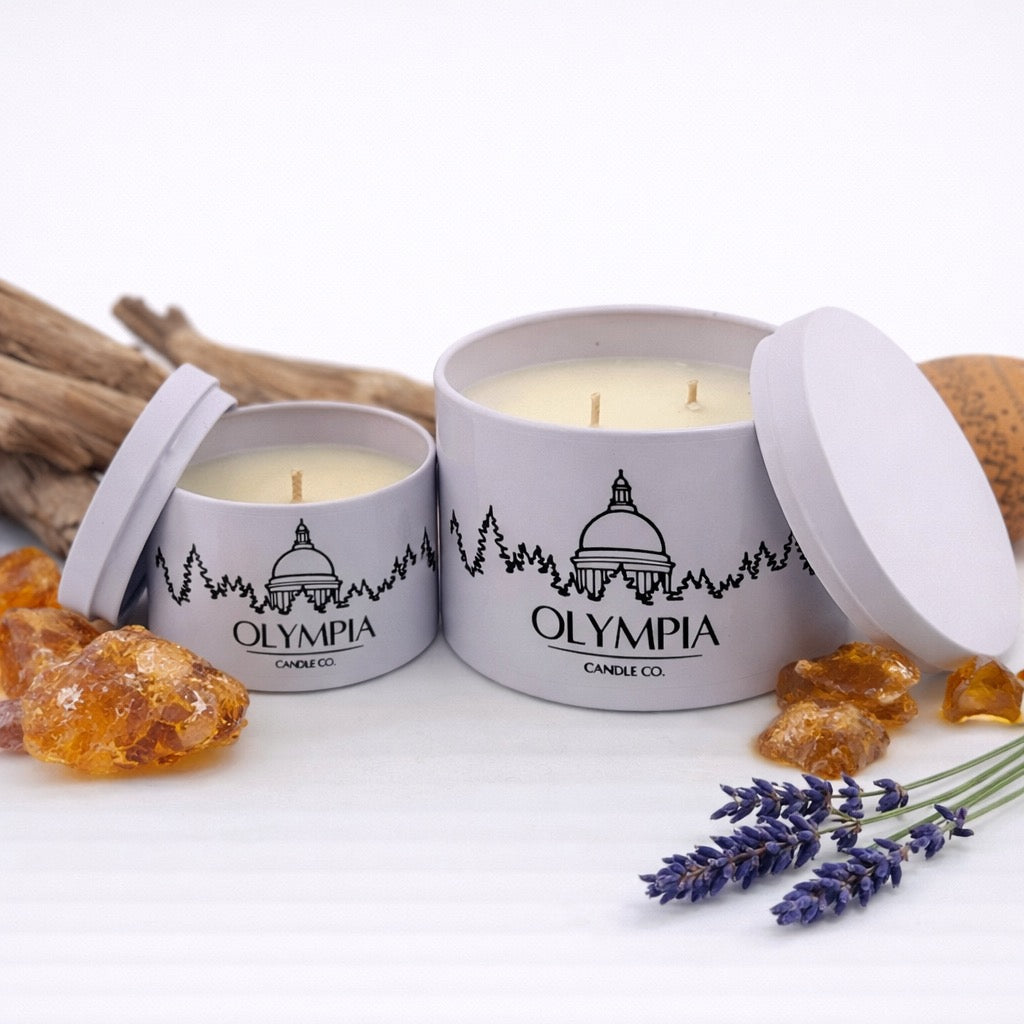 Two Olympia Candle Co. candles in white tins with a decorative lid, surrounded by amber gummy bears and lavender flowers on a white background.