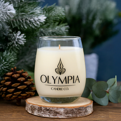 Glass candle with Olympia Candle Co. logo on a wooden coaster with greenery in the background