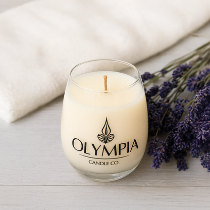 White candle in a glass jar with 'Olympia Candle Co.' logo, surrounded by lavender flowers on a light surface.
