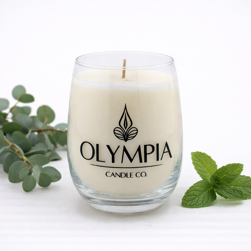 Glass candle with 'Olympia Candle Co.' label surrounded by green leaves on a white background