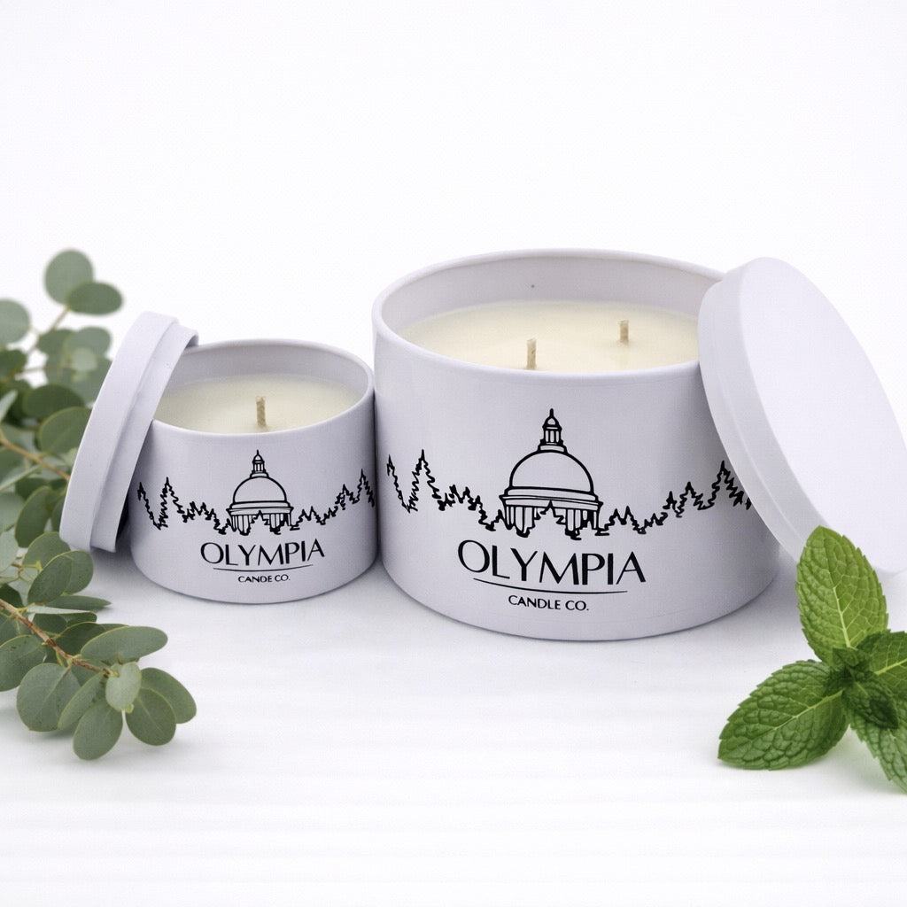 Two candles in white tins with 'Olympia Candle Co' branding, surrounded by green leaves on a white background.