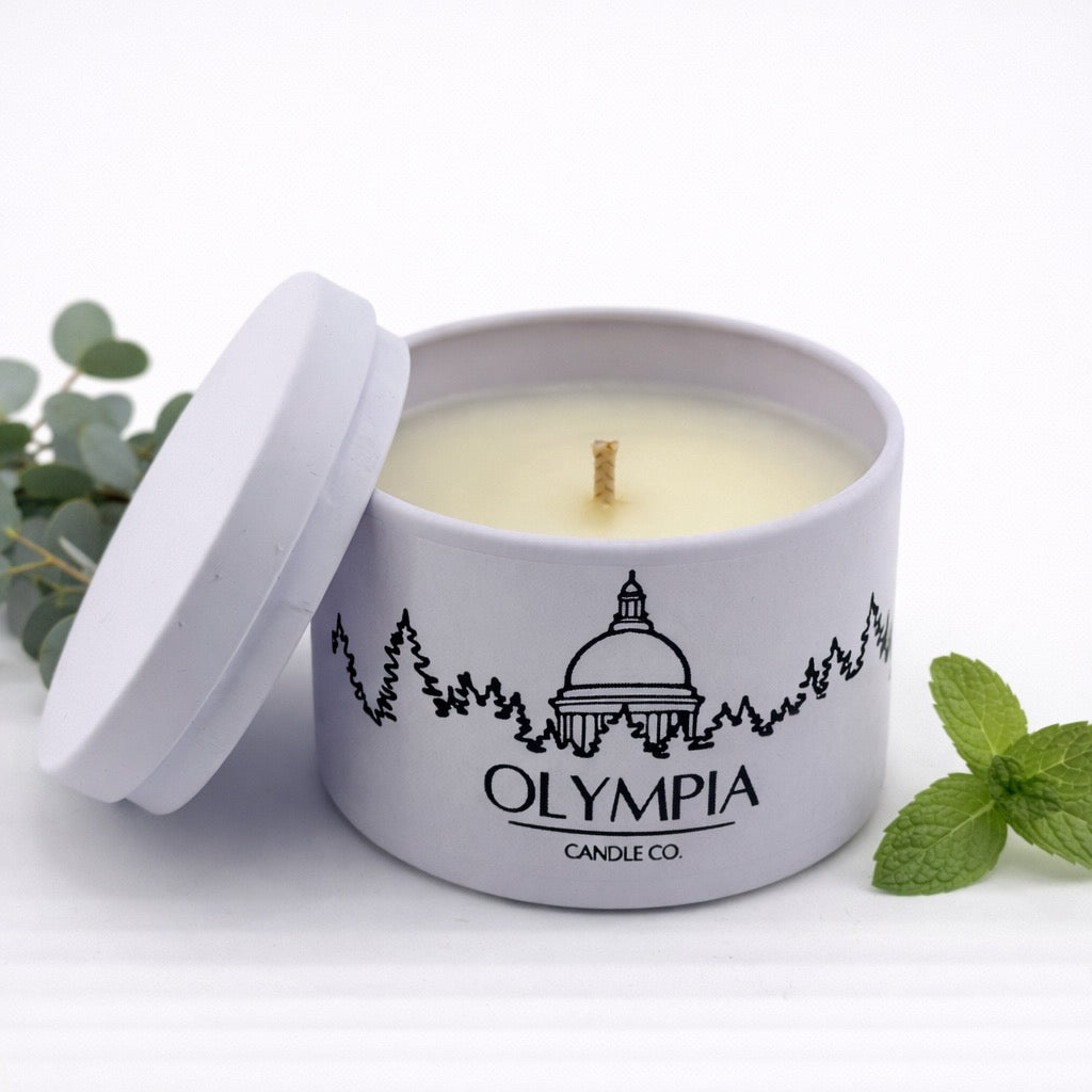 Candle in a tin with 'Olympia Candle Co.' branding, surrounded by greenery on a white background