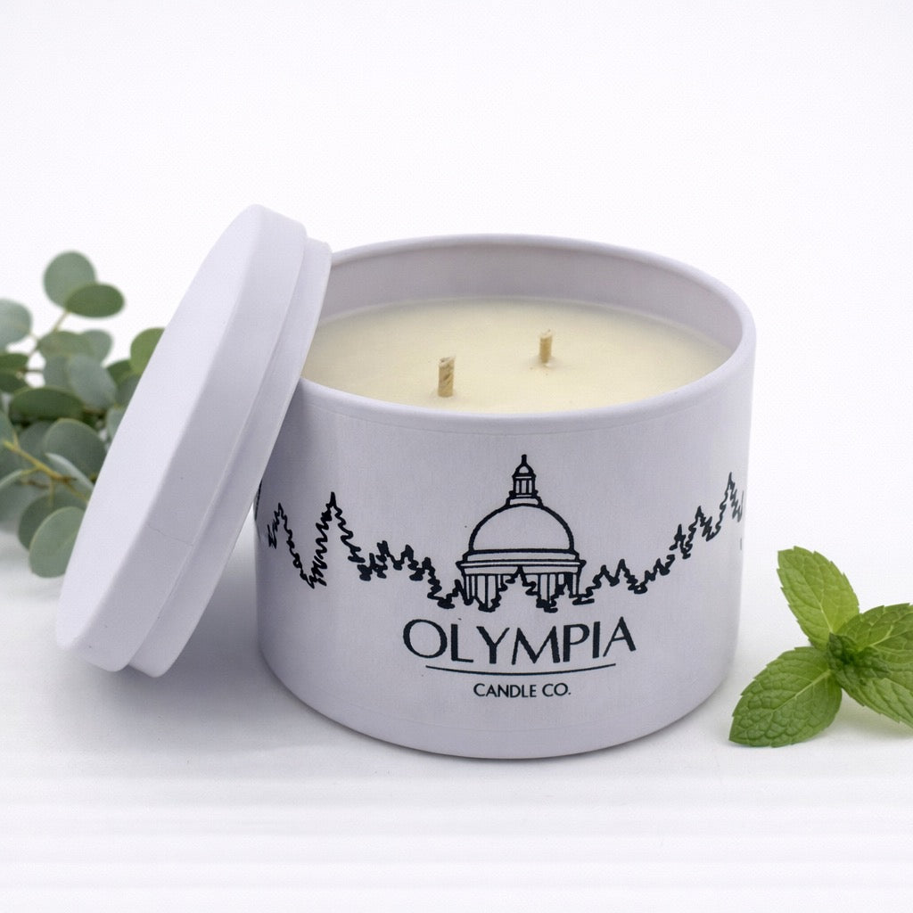 Olympia Candle Co. candle in a gray tin with eucalyptus and mint leaves on a white background