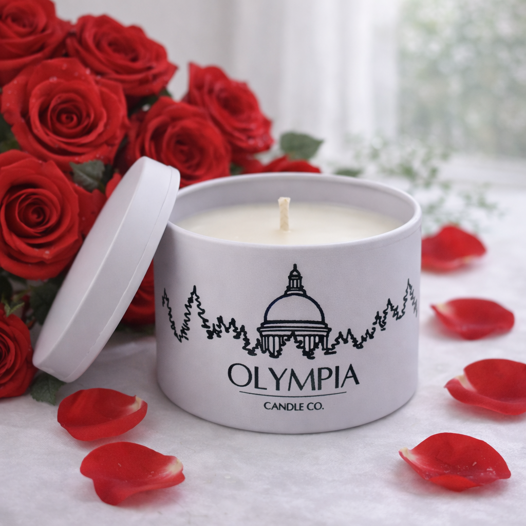 White candle in a container with 'Olympia Candle Co.' logo, surrounded by red roses and petals.