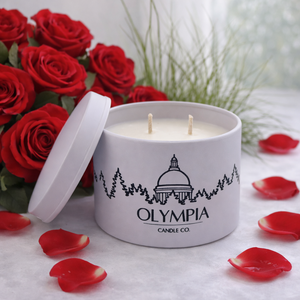 White candle with 'Olympia Candle Co.' logo surrounded by red roses and petals on a light surface.